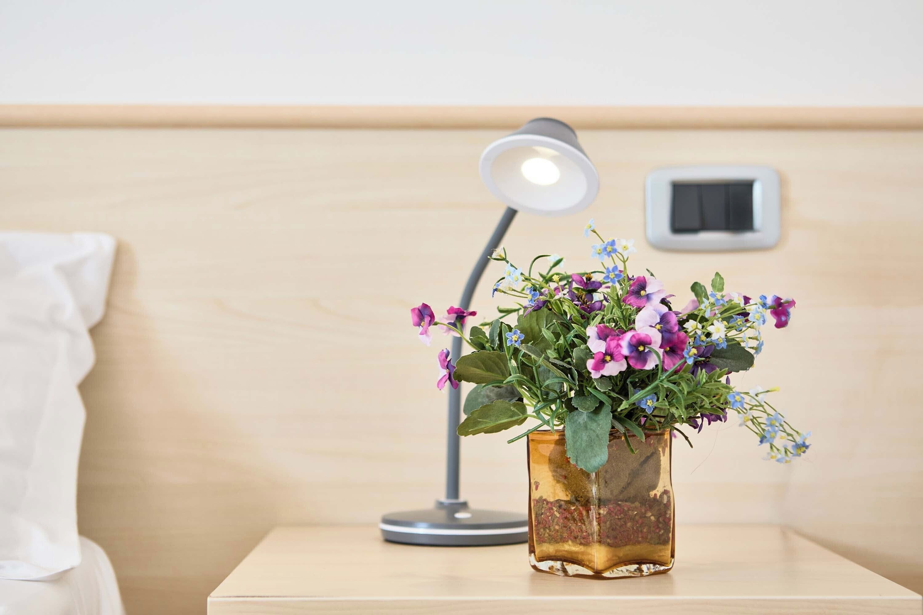 Een nachtkastje met een glazen vaas met bloemen, een lamp en een stopcontact op een houten ondertafel.