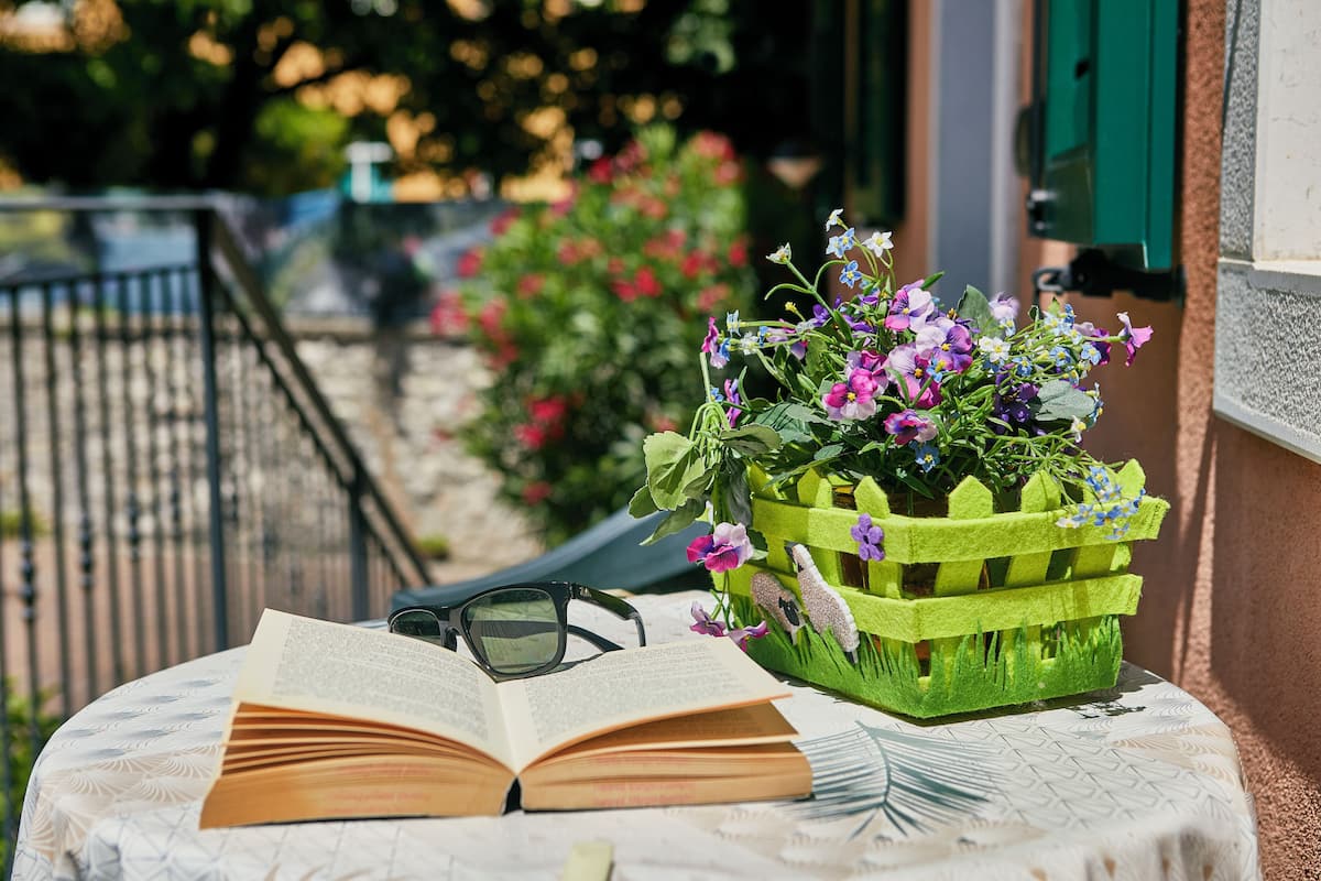 An open book on a table with a pair of glasses beside it, a green basket with flowers, and a railing in the background.