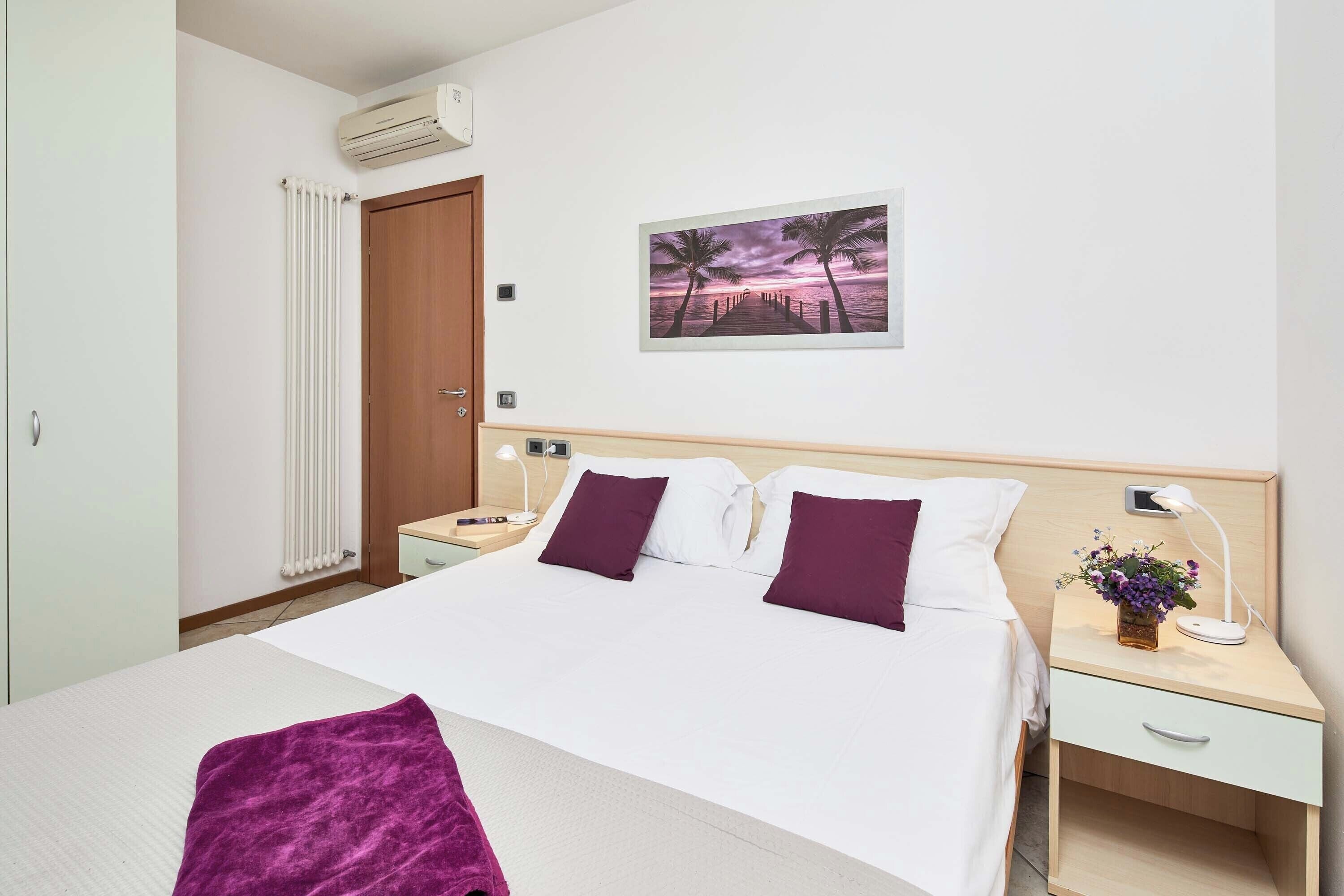 Et hotelværelse med en seng, et natbord, en lampe på natbordet, en fastgjort aircondition, en radiator og et billede i ramme på væggen.