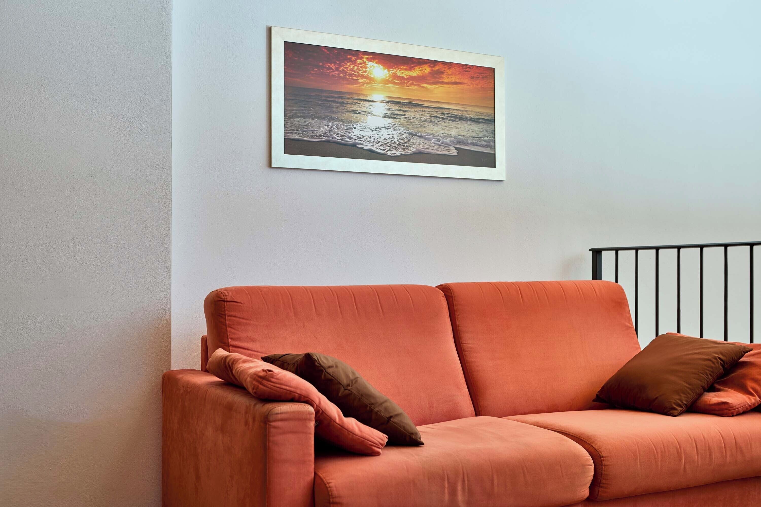 A room with a single orange sofa against a wall, a framed picture of a sunset over the ocean, and a black metal railing.