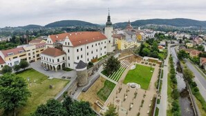 Vista aérea de uma cidade histórica com um castelo imponente, uma igreja e uma praça pública bem cuidada com uma fonte.