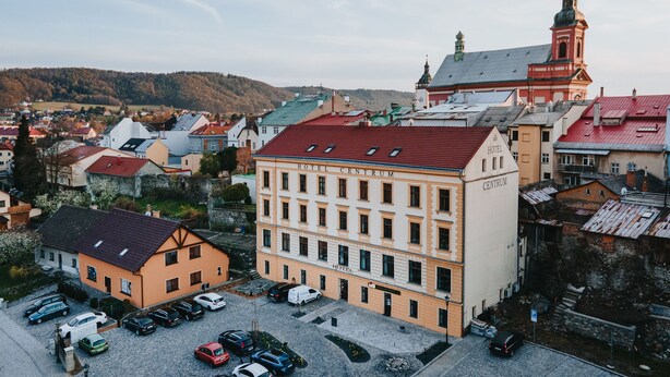 Um hotel com telhado vermelho e uma torre do relógio, cercado por outros edifícios e uma paisagem montanhosa ao fundo.