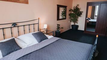 A neatly made bed with a grey quilt, a bedside lamp, a potted plant, and a mirror on the wall.