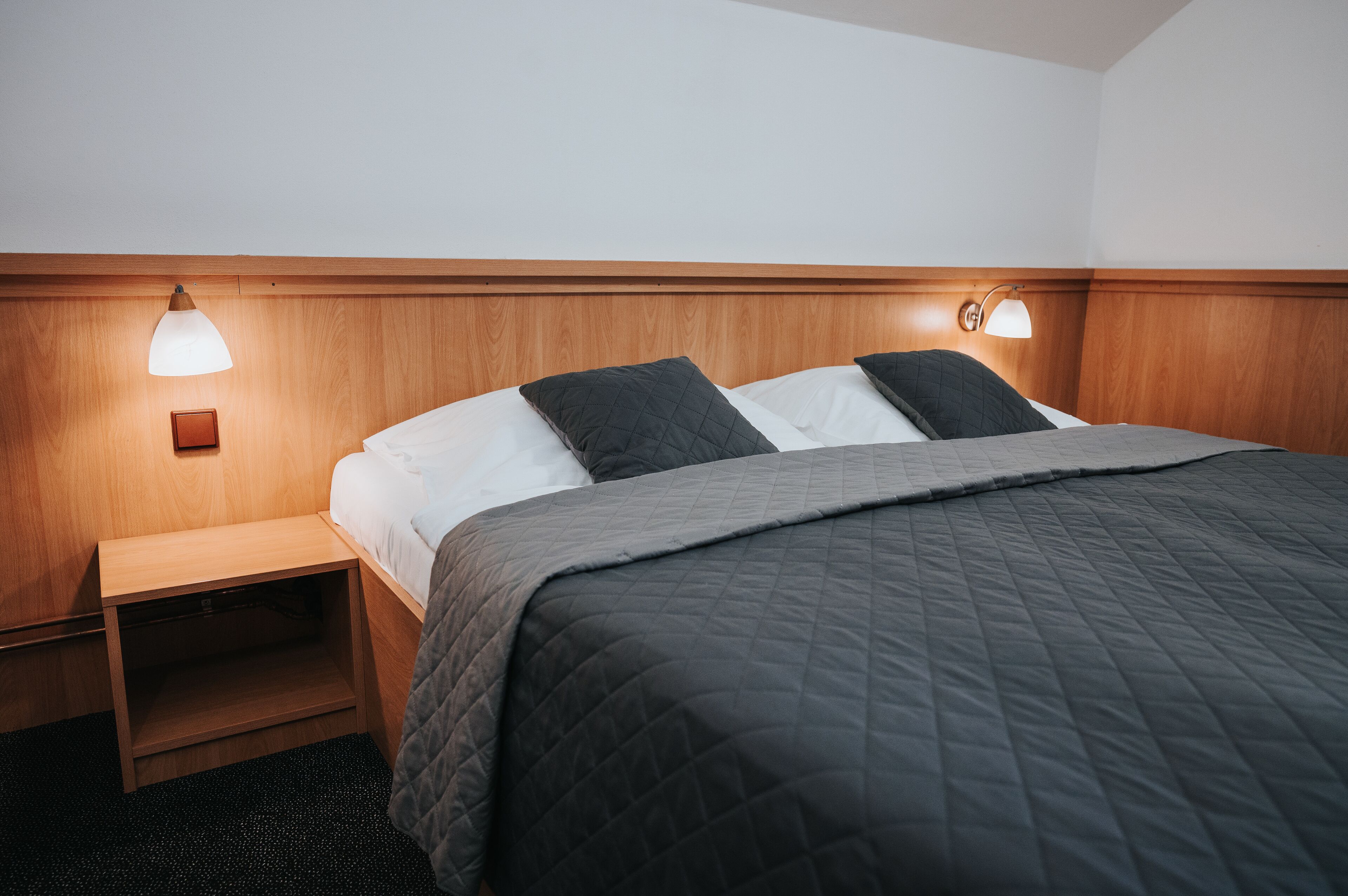 A neatly made bed with a dark quilt, a wooden headboard, and two bedside lamps.