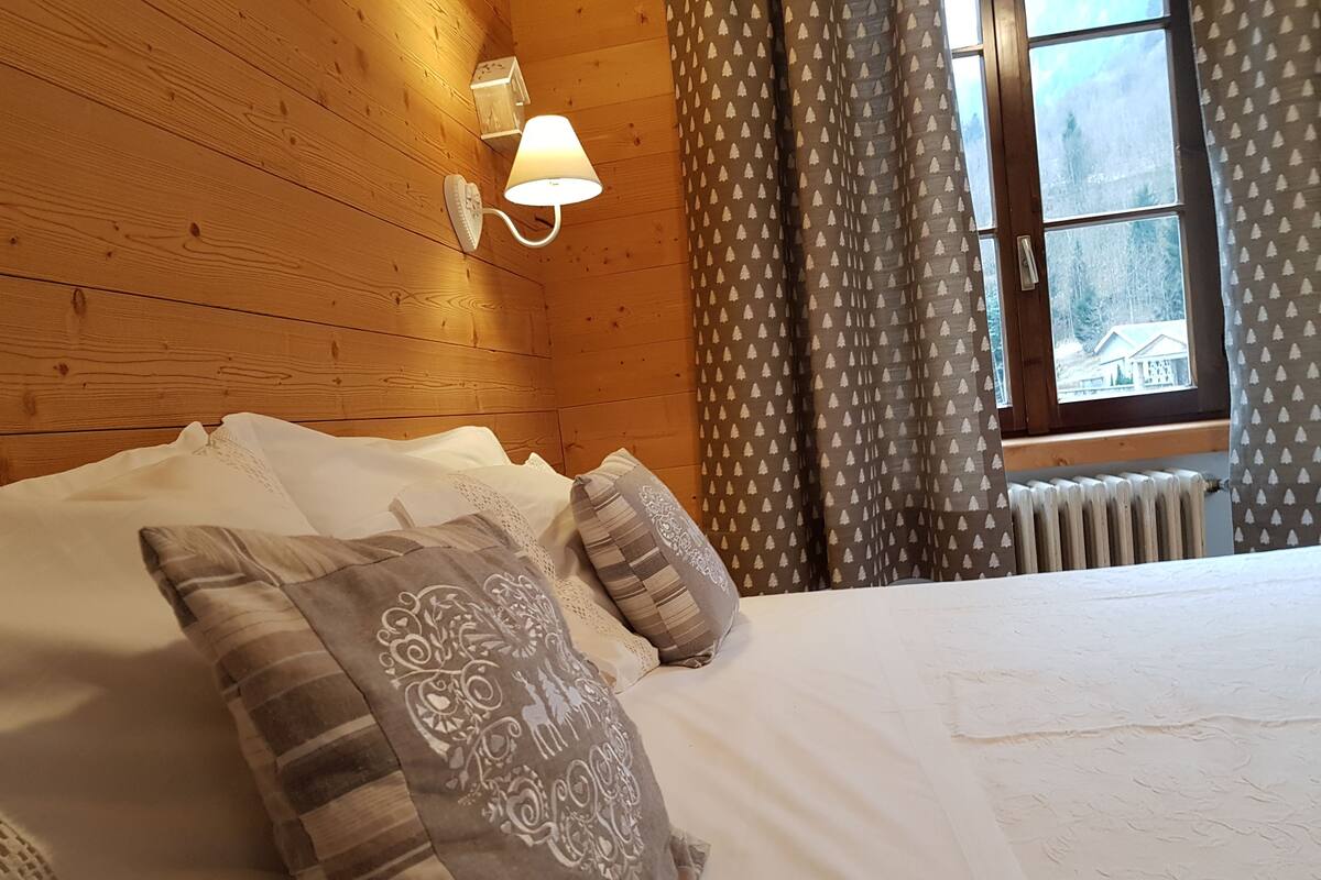A wooden-paneled room with a bed, decorative pillows, a wall-mounted lamp, and a window with curtains.