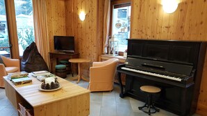A cozy room with a wooden interior, a piano, a television, and a dining table with books and a candle. - Hotel Pedretti (Branzi)