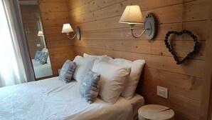 A neatly made bed with white linens and patterned pillows, a wooden headboard, a wall-mounted lamp, and a heart-shaped decoration on the wall. - Hotel Pedretti (Branzi)
