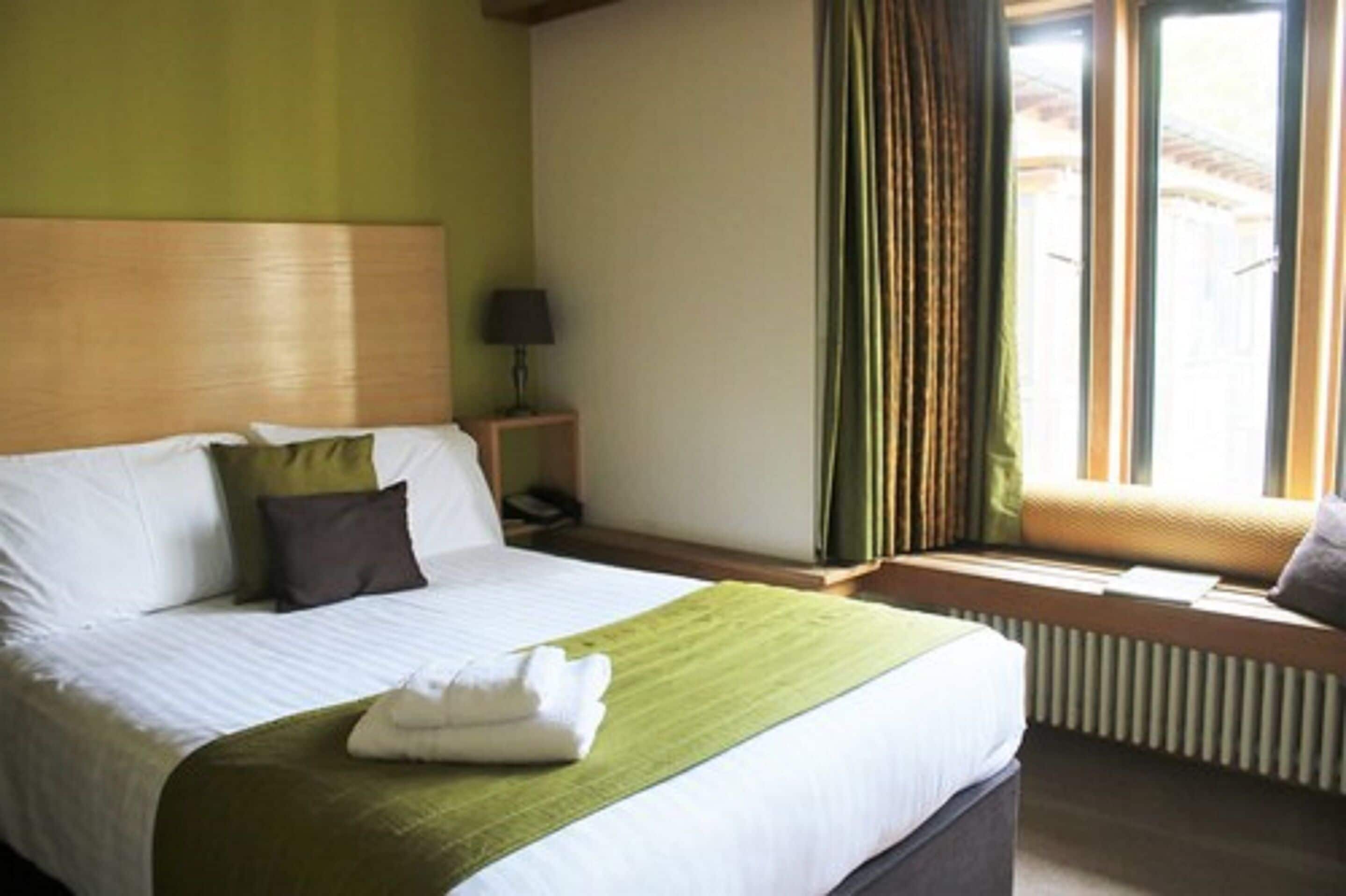 Une chambre d’hôtel avec un lit, une table de chevet avec une lampe, une fenêtre avec des rideaux et un radiateur.