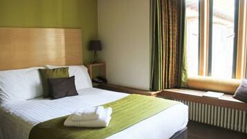A hotel room with a bed, a bedside table with a lamp, a window with curtains, and a radiator.