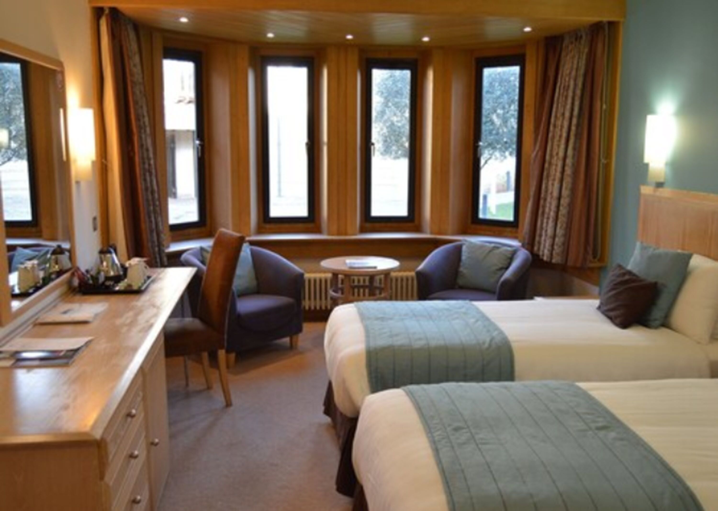 Habitación de hotel con cama, dos sillones, un escritorio con lámpara y ventanas grandes con cortinas.