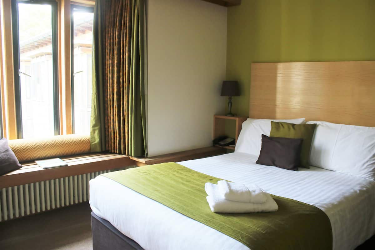 A hotel room with a large bed, a window with curtains, a bedside table with a lamp, and a radiator.