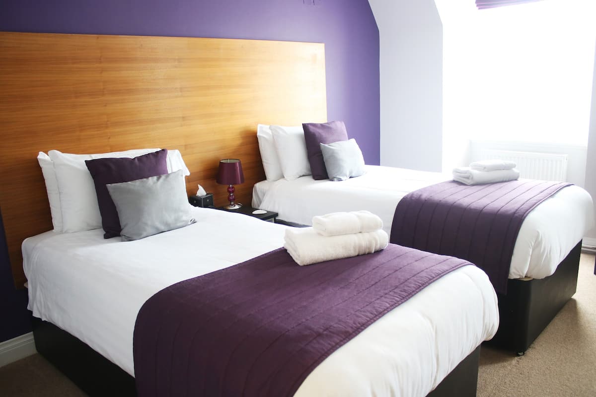 A hotel room with two beds, each with a wooden headboard, white bedding, and purple accents. There is a nightstand with a lamp and a folded towel on each bed.