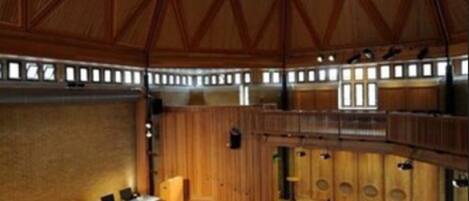 Un salón amplio con techo de madera y un escenario central. Filas de sillas están dispuestas para el público.