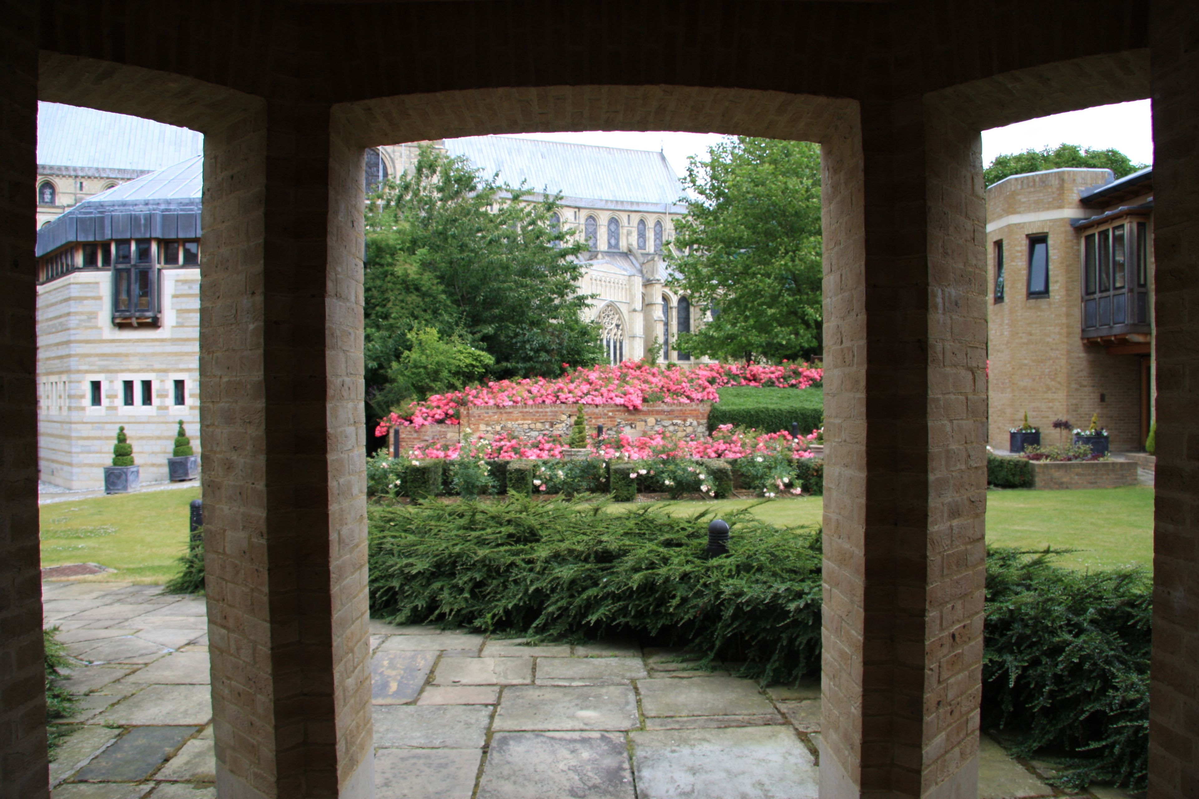 A stone archway framing a view of a historic building with a garden of pink flowers.