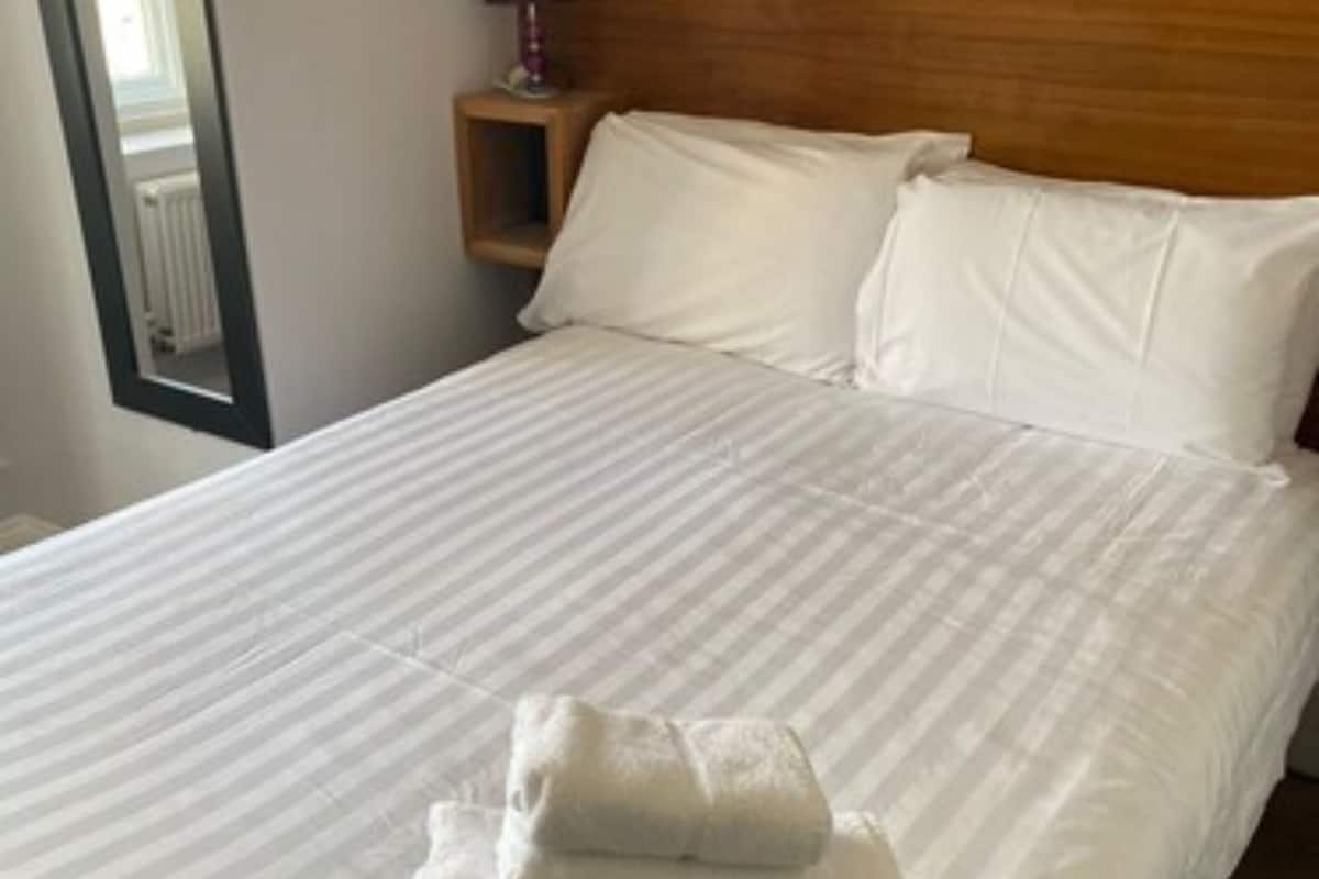 A neatly made bed with white linens and two pillows, a wooden headboard, a nightstand with a lamp, and a window with blinds.