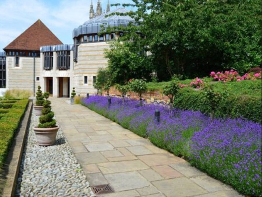 A building with a central entrance, surrounded by a garden with purple flowers and neatly trimmed hedges.
