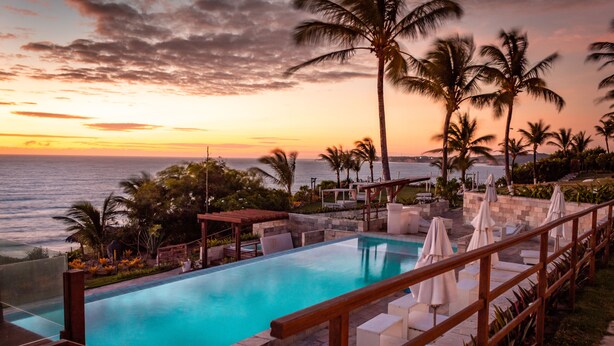 Un área de piscina con muelle de madera, sillones de descanso y sombrillas, con vista al océano al atardecer.