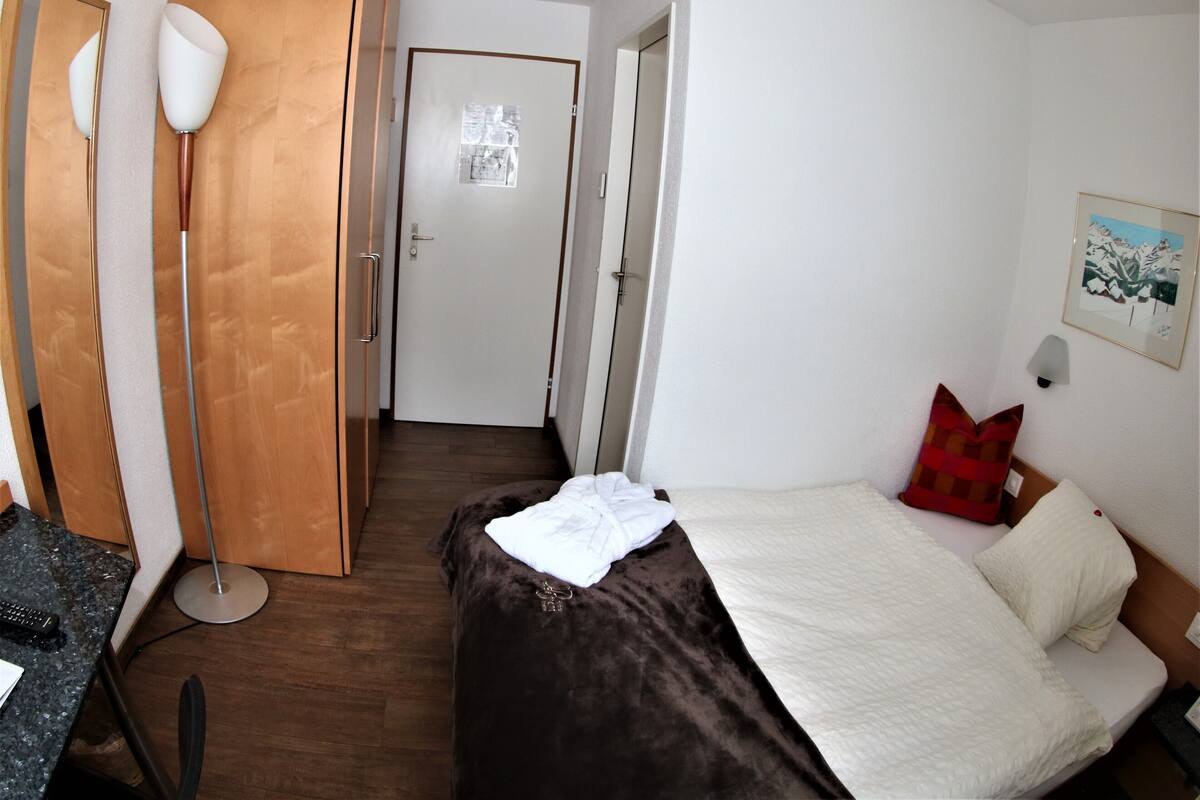 Une chambre d’hôtel avec un lit, un bureau, une chaise, une lampe sur pied et une porte.