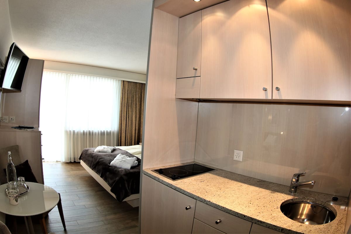 Une chambre d’hôtel compacte comprenant un coin cuisine, un lit recouvert d’une couverture foncée, une petite table avec une bouteille et des verres, et un téléviseur fixé au mur.