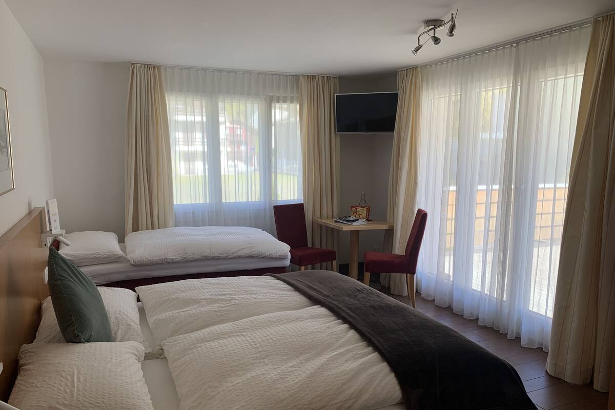 Une chambre d’hôtel avec deux lits, une télévision, une petite table avec une chaise et de grandes fenêtres avec des rideaux.