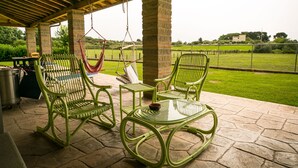 Un porche cubierto con mobiliario de mimbre verde, incluyendo dos mecedoras y una mesa, con vistas a un campo cubierto de césped.