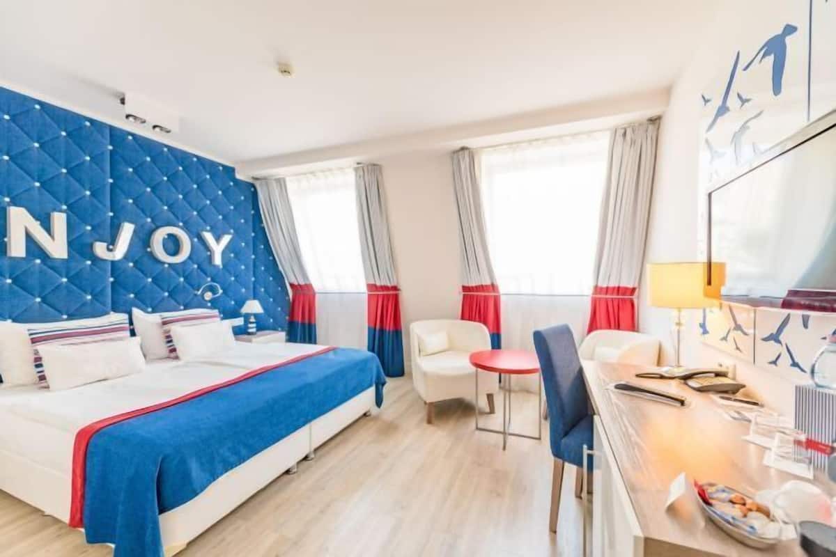 A hotel room with a bed, a desk, a chair, a window with curtains, and a wall with the word 'ENJOY'.