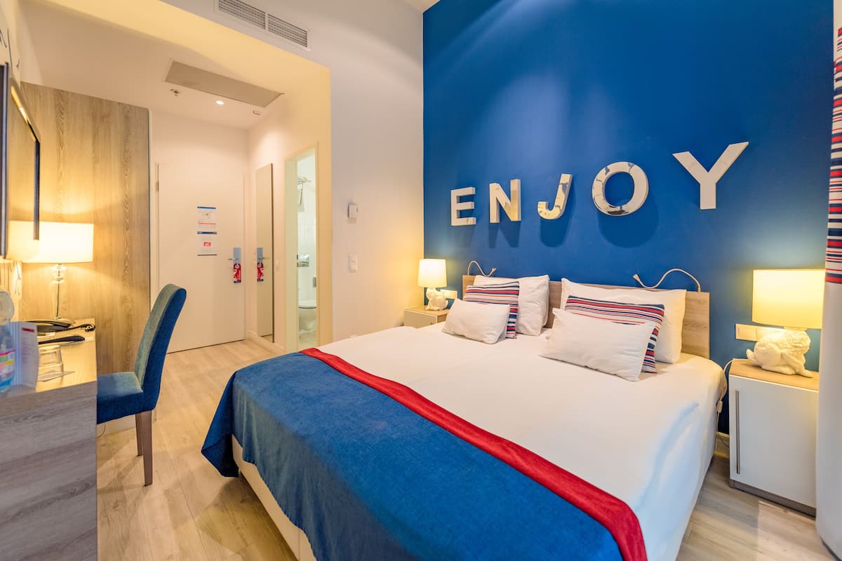 A hotel room with a bed, a desk, and a chair. There is a wall with the word "ENJOY" on it.