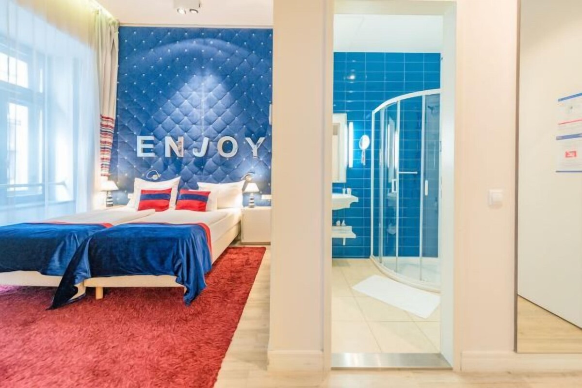 Un dormitorio con una cama, una alfombra roja, una pared azul con el texto 'ENJOY' y un baño con azulejos azules y una ducha de vidrio.