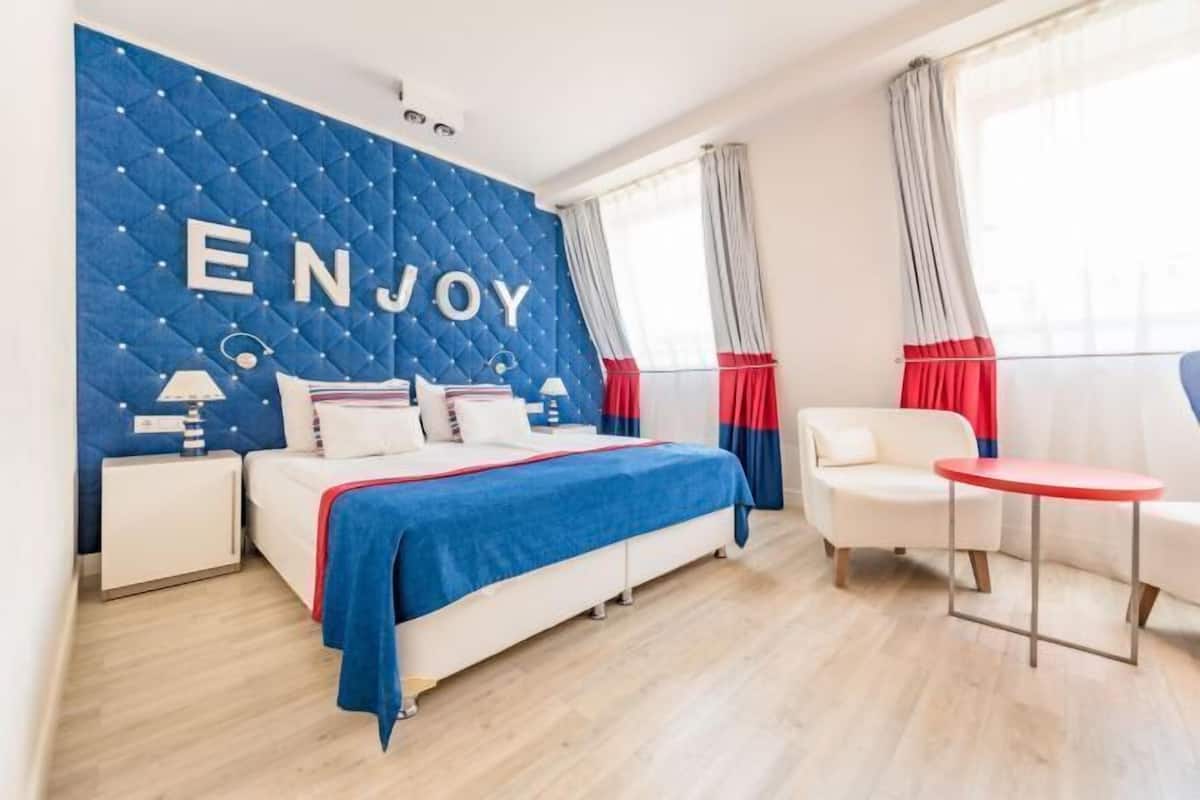 A modern hotel room with a bed, a small table, a chair, a nightstand, and a wall with the word 'ENJOY'.