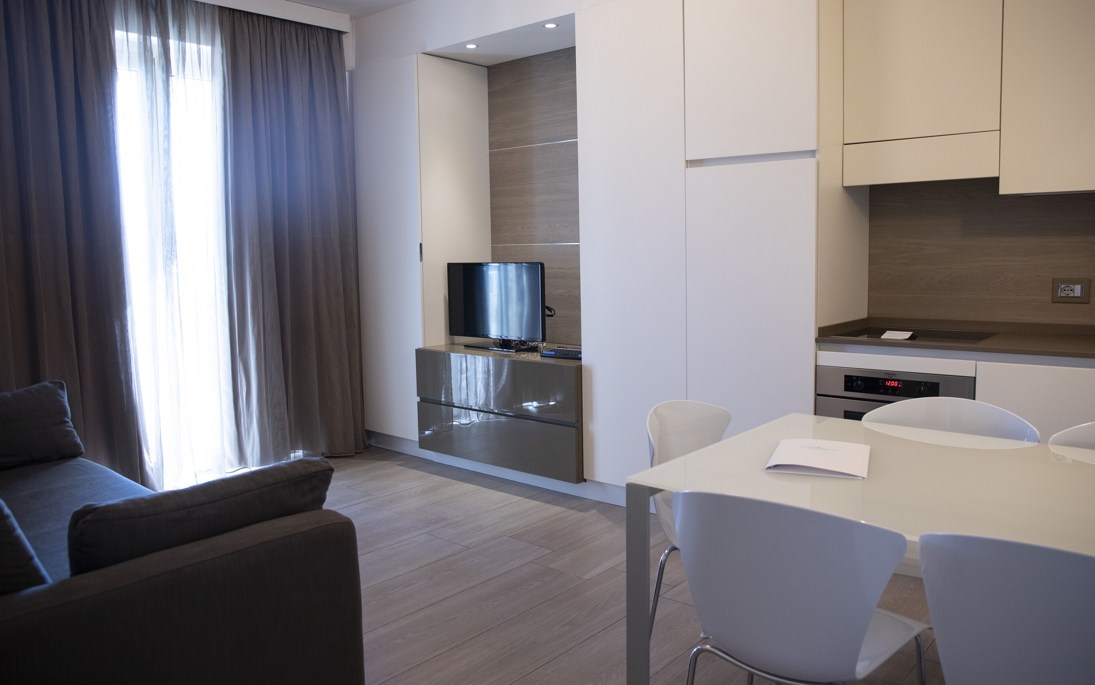 Ein modernes Hotelzimmer mit einer Couch, einem Esstisch mit Stühlen, einem Fernseher, einer Küchenzeile mit Herd und einem großen Fenster mit Vorhängen.