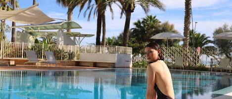 Un área de piscina con sillas de descanso y sombrillas, rodeada de palmeras.