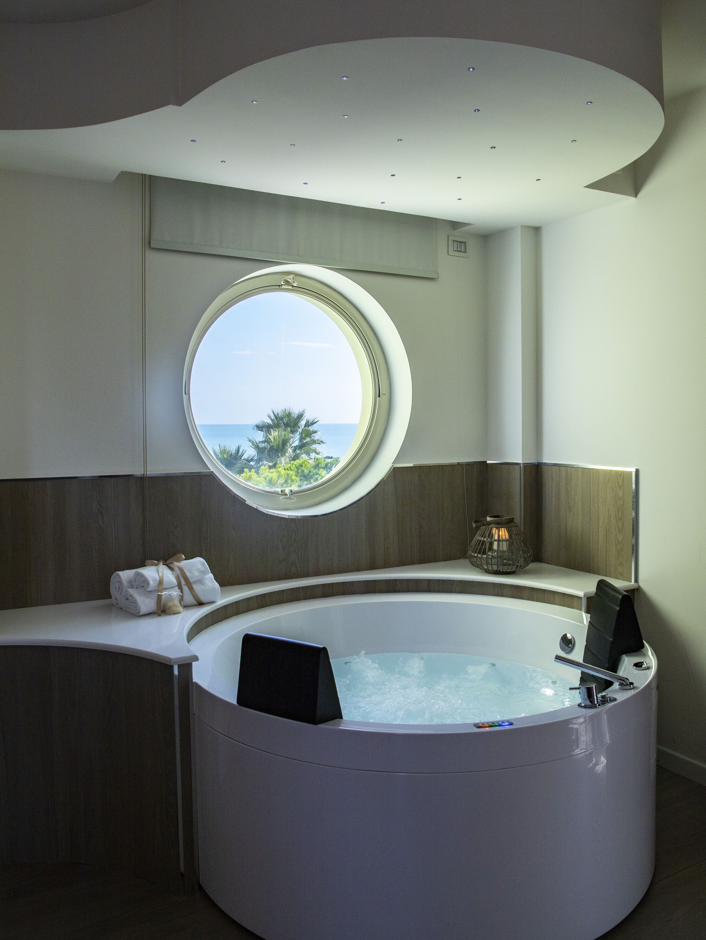 A modern bathroom with a large bathtub, a round window offering a view of the sea, and a small table with a candle.