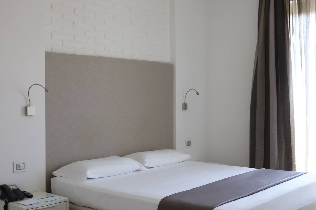 A modern hotel room with a single bed, a bedside table, a lamp, and a window with curtains.