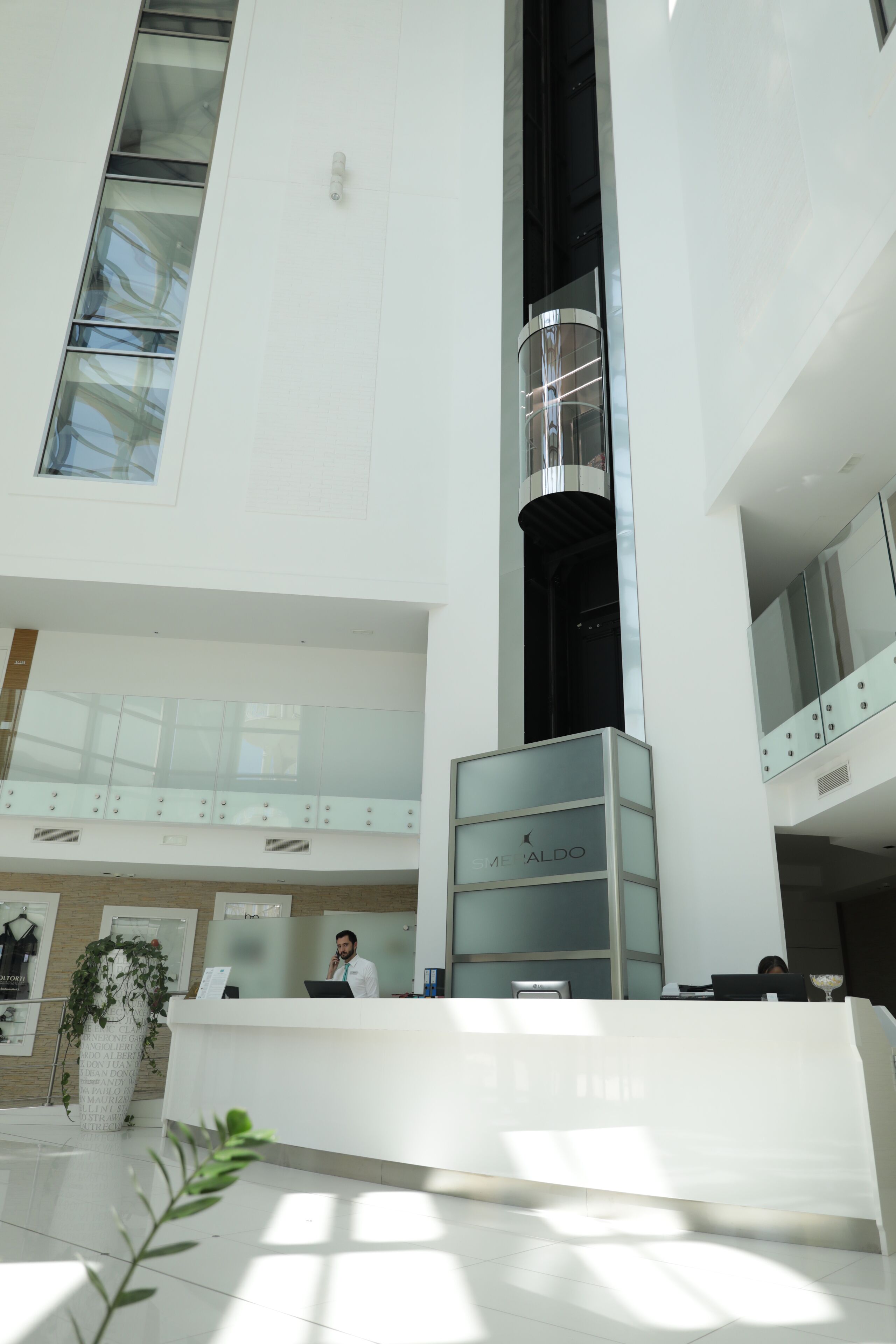 A modern hotel lobby with a reception desk, a vertical elevator shaft, and a glass railing on the upper level.