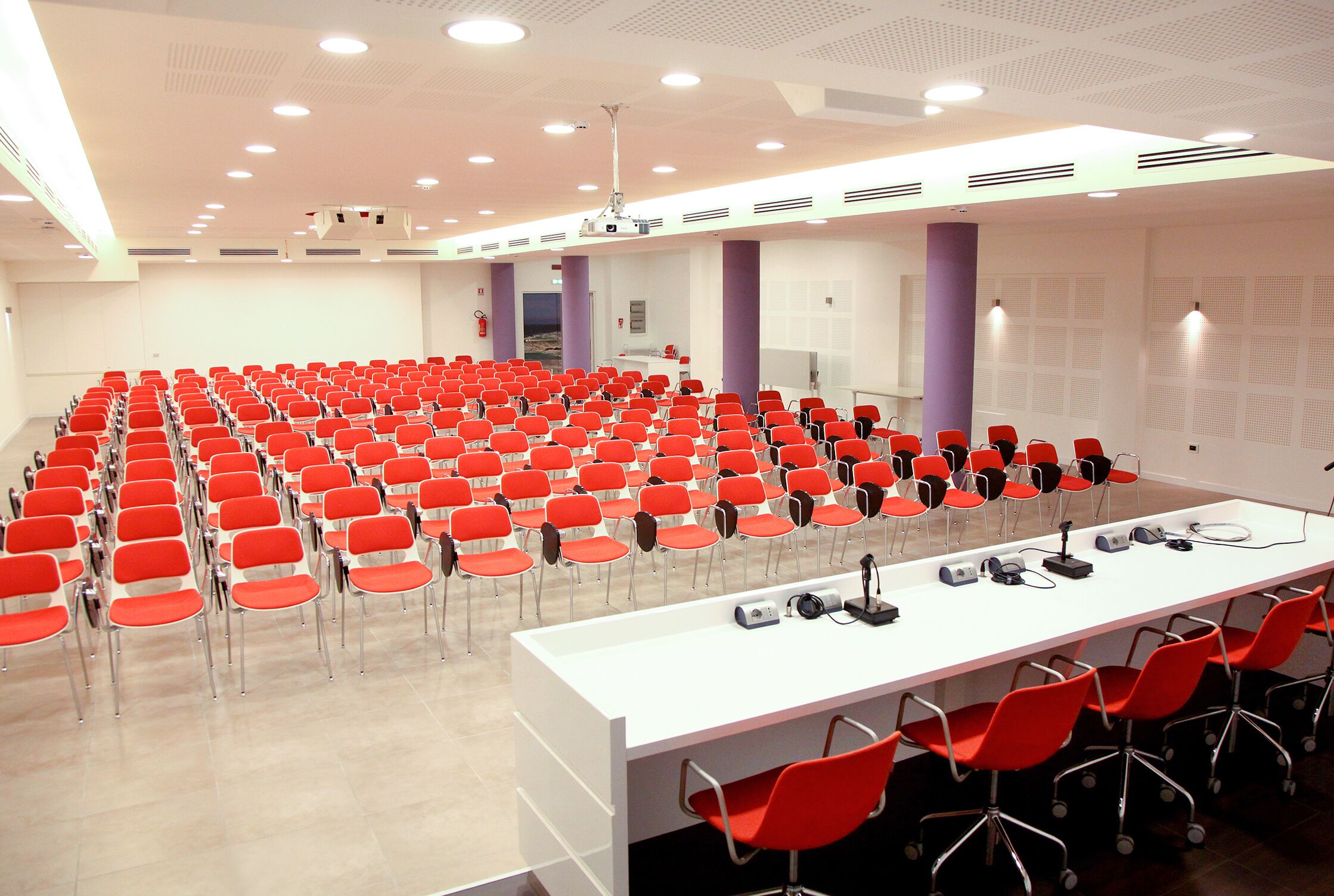 A conference room with rows of red chairs, a white table with microphones, and a projector screen.