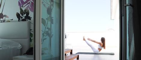 A room with a bed, a nightstand, and a window with a view of a balcony where a person is doing yoga.