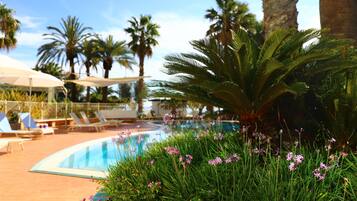 Un área de piscina con sillones de descanso y sombrillas, rodeada de palmeras y plantas con flores.