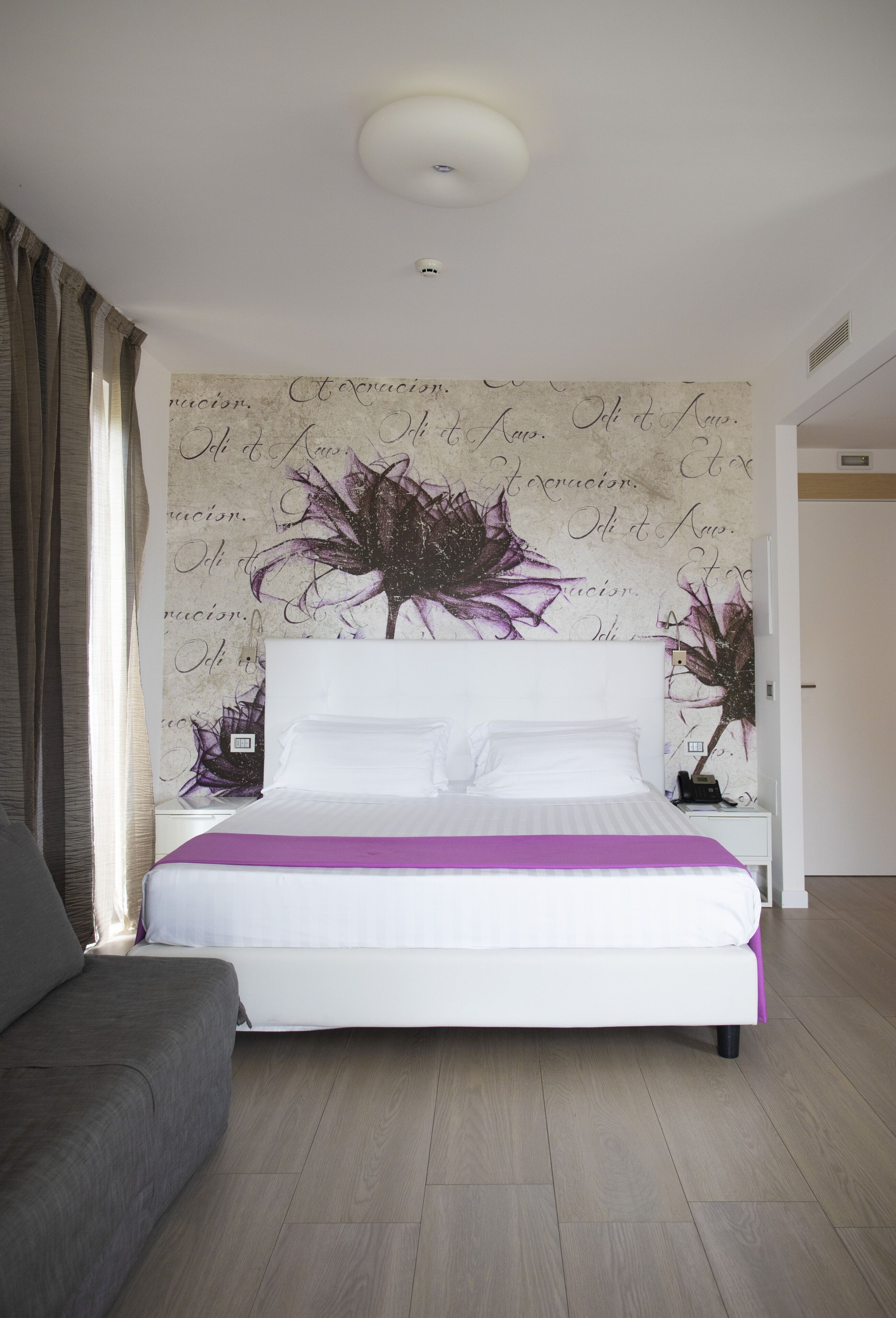 A bedroom with a bed, a nightstand, a grey sofa, and a wall with floral wallpaper.
