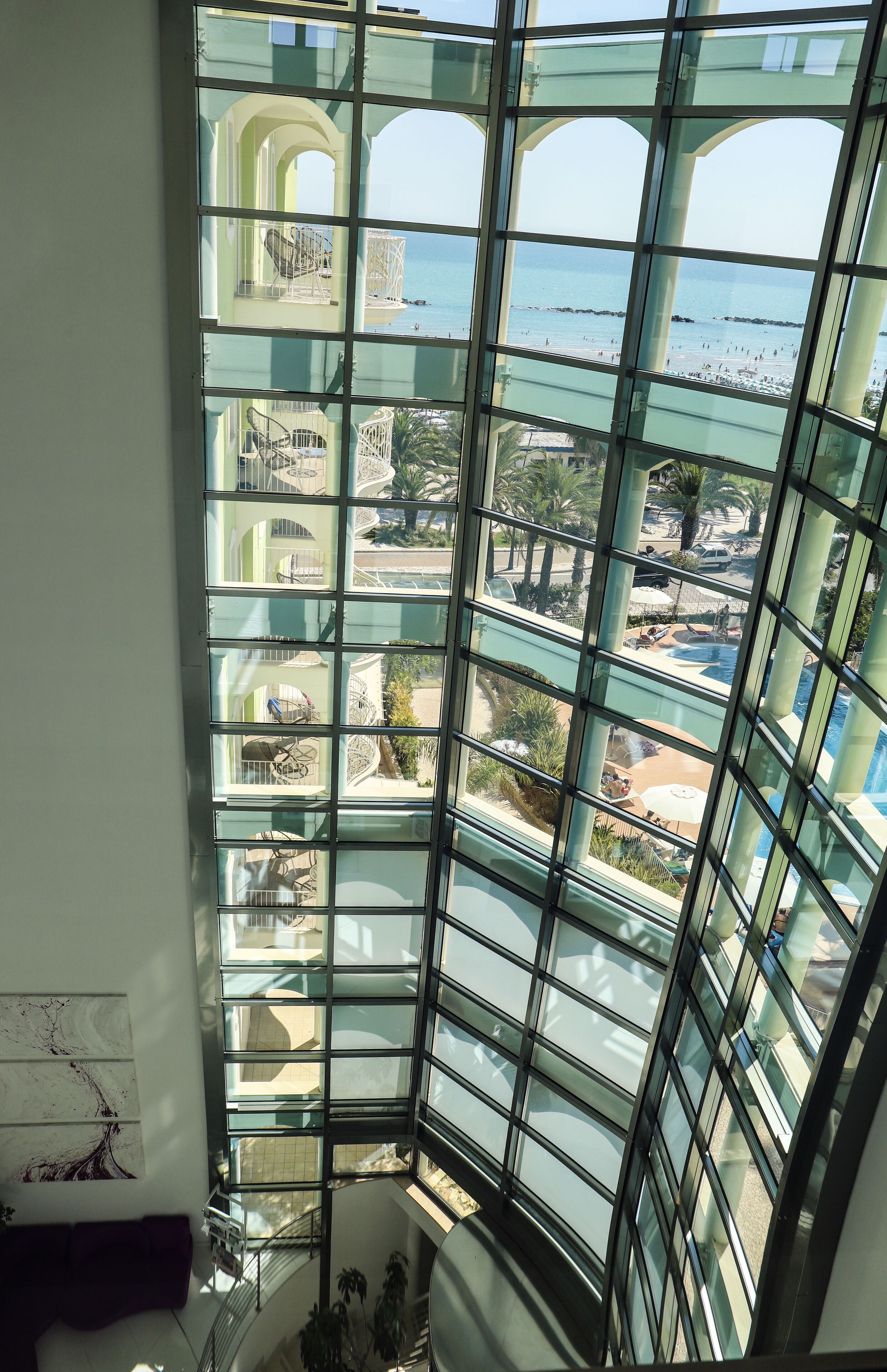 A high-rise hotel interior with a large glass window offering a view of a pool, lounge area, and beach.