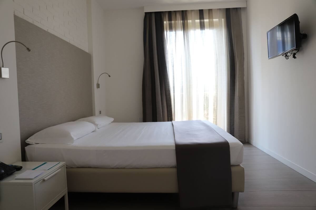 A neatly made bed with white linens, a bedside table with a lamp, and a flat-screen TV mounted on the wall.