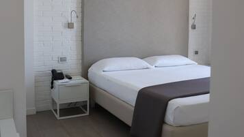 A hotel room with a bed, a nightstand with a phone and a lamp, and a white brick wall.
