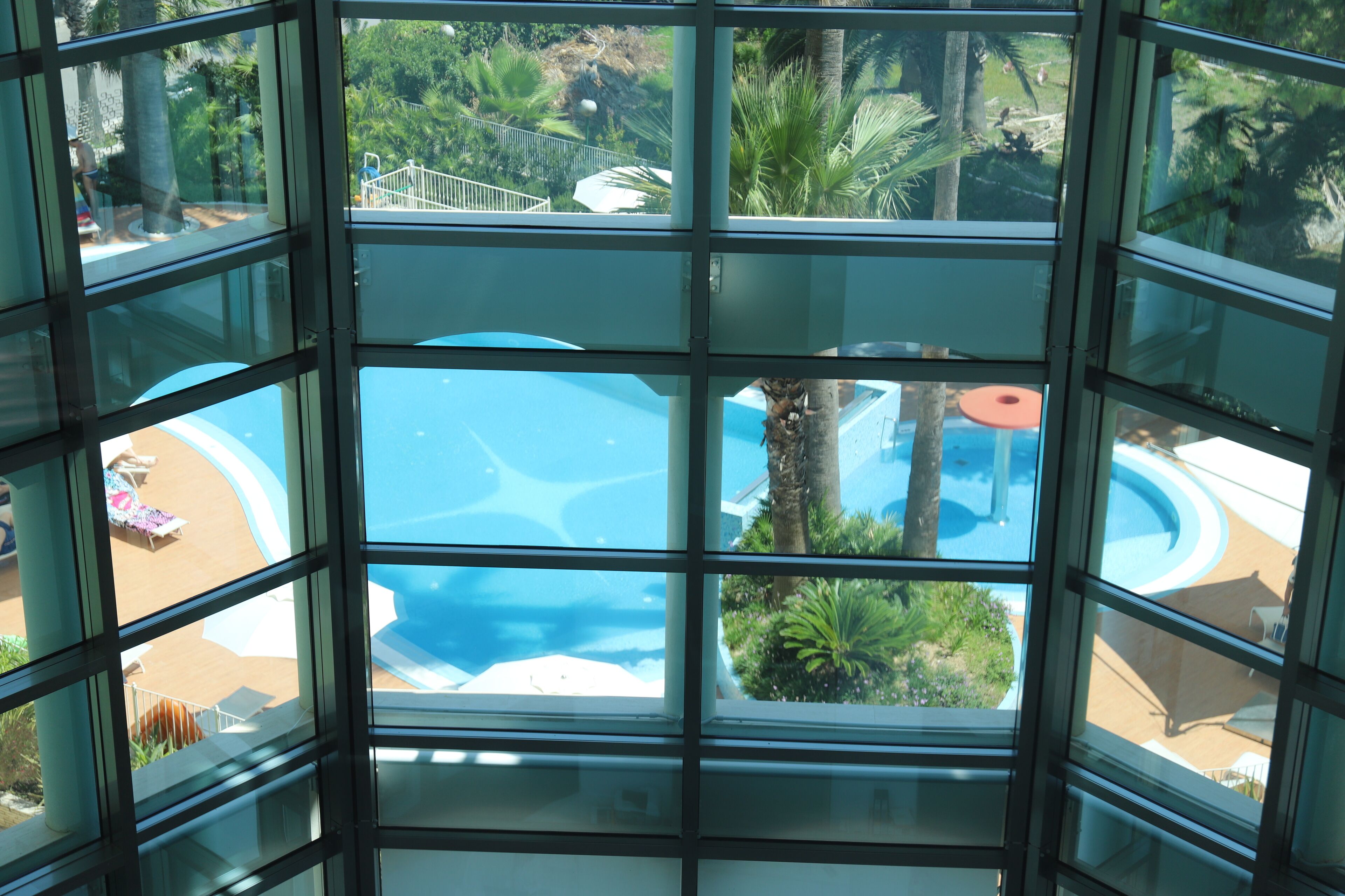 Vista a través de un ventanal de un área de piscina con una fuente circular, sillones de descanso y palmeras.