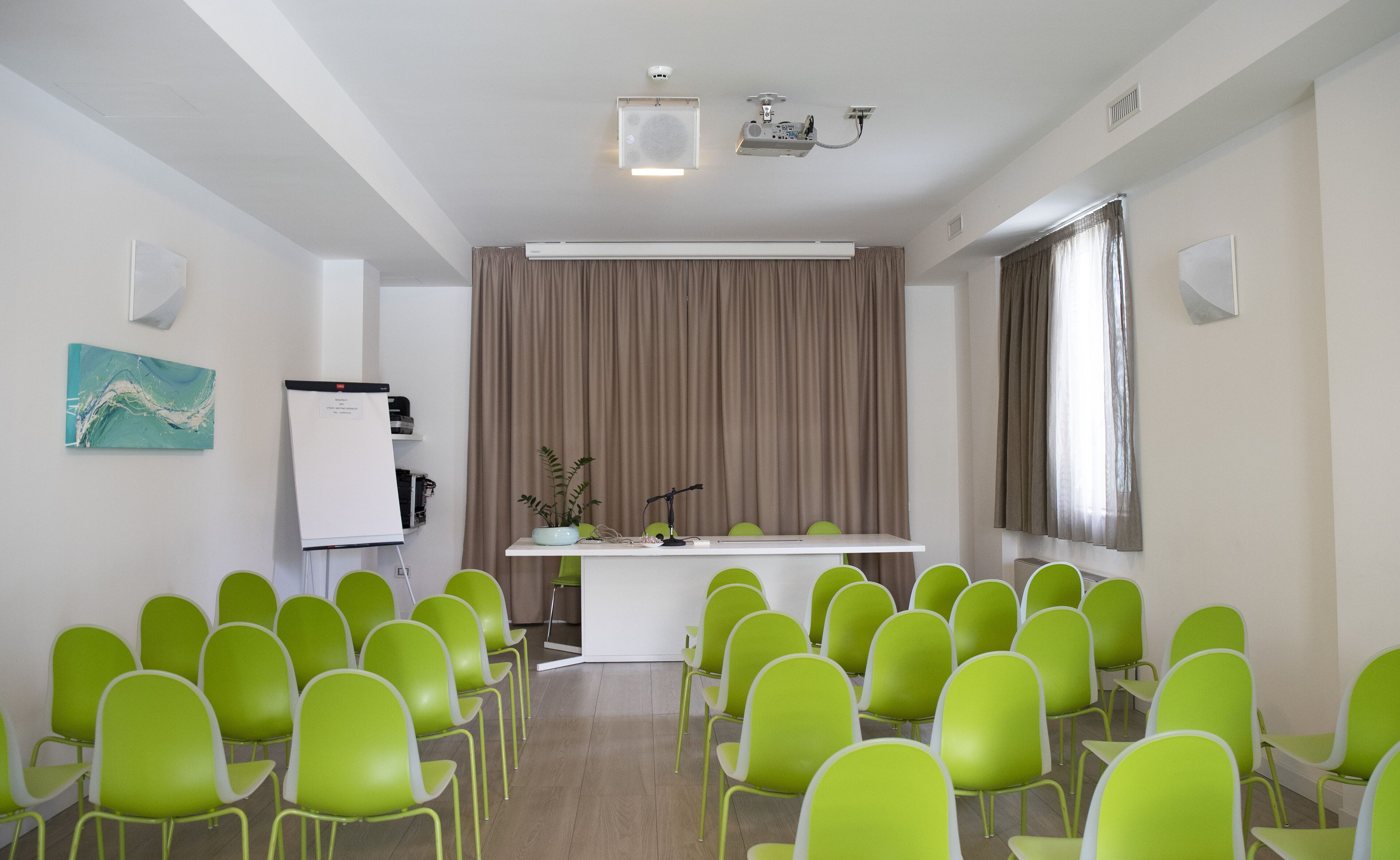 Un salón de conferencias con sillas verdes dispuestas en filas, una pizarra y un proyector sobre un soporte.