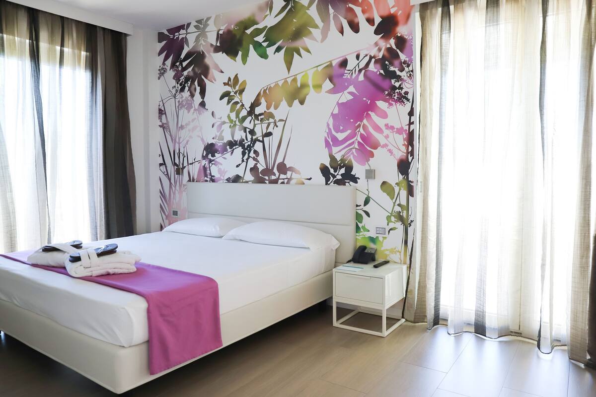 A neatly made bed with a pink blanket, a white bedside table with a phone and a lamp, and a wall with a floral mural.