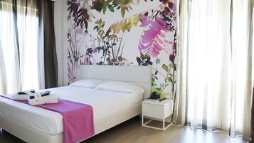 A neatly made bed with a pink blanket, a white bedside table with a phone and a lamp, and a wall with a floral mural.