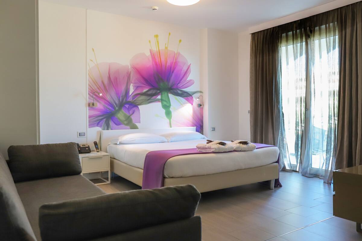 A modern hotel room with a bed, a sofa, a nightstand, and a large window with curtains.