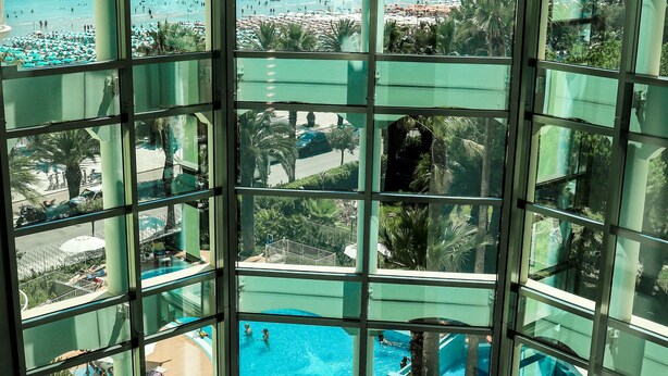 A glass-enclosed area with a pool, palm trees, and a view of the beach. - Smeraldo Suites & Spa (San Benedetto del Tronto)