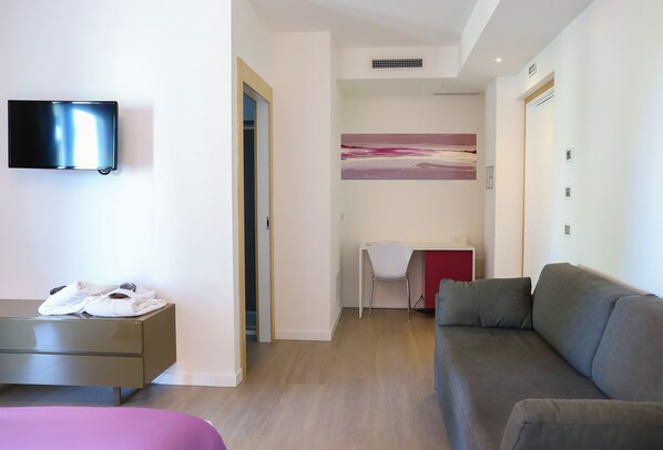 A modern hotel room with a flat-screen TV, a desk with a chair, a grey sofa, and a painting on the wall.