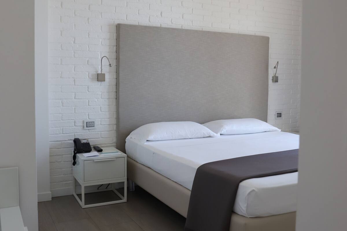 A modern hotel room with a white brick wall, a large bed with a grey headboard, a bedside table with a lamp, and a nightstand with a phone.