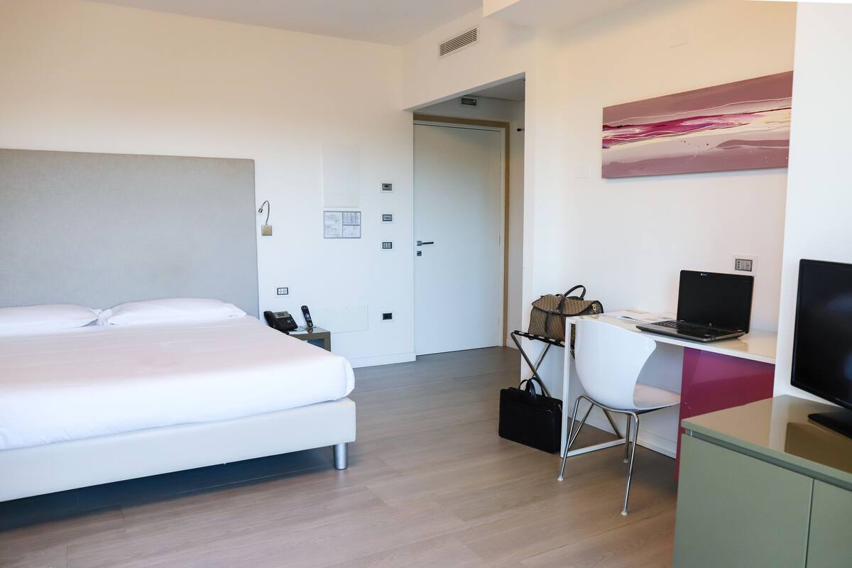 A modern hotel room with a bed, a desk with a laptop, a chair, a TV, and a painting on the wall.